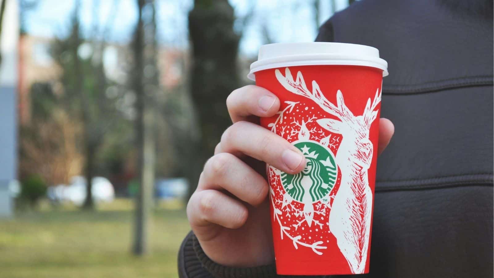 Person holding a red Starbucks holiday cup with a white deer design outdoors, enjoying one of the festive Starbucks Holiday Drinks&mdash;perfect for sipping hot or iced in the chilly air.