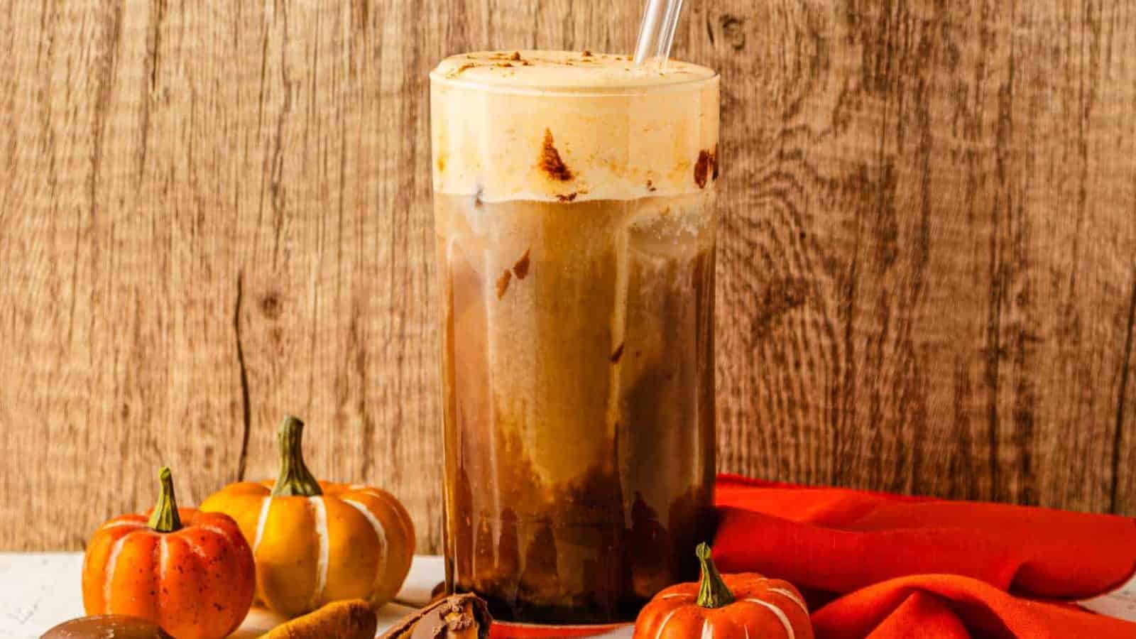Iced coffee with creamy foam in a tall glass, surrounded by small pumpkins and an orange cloth.