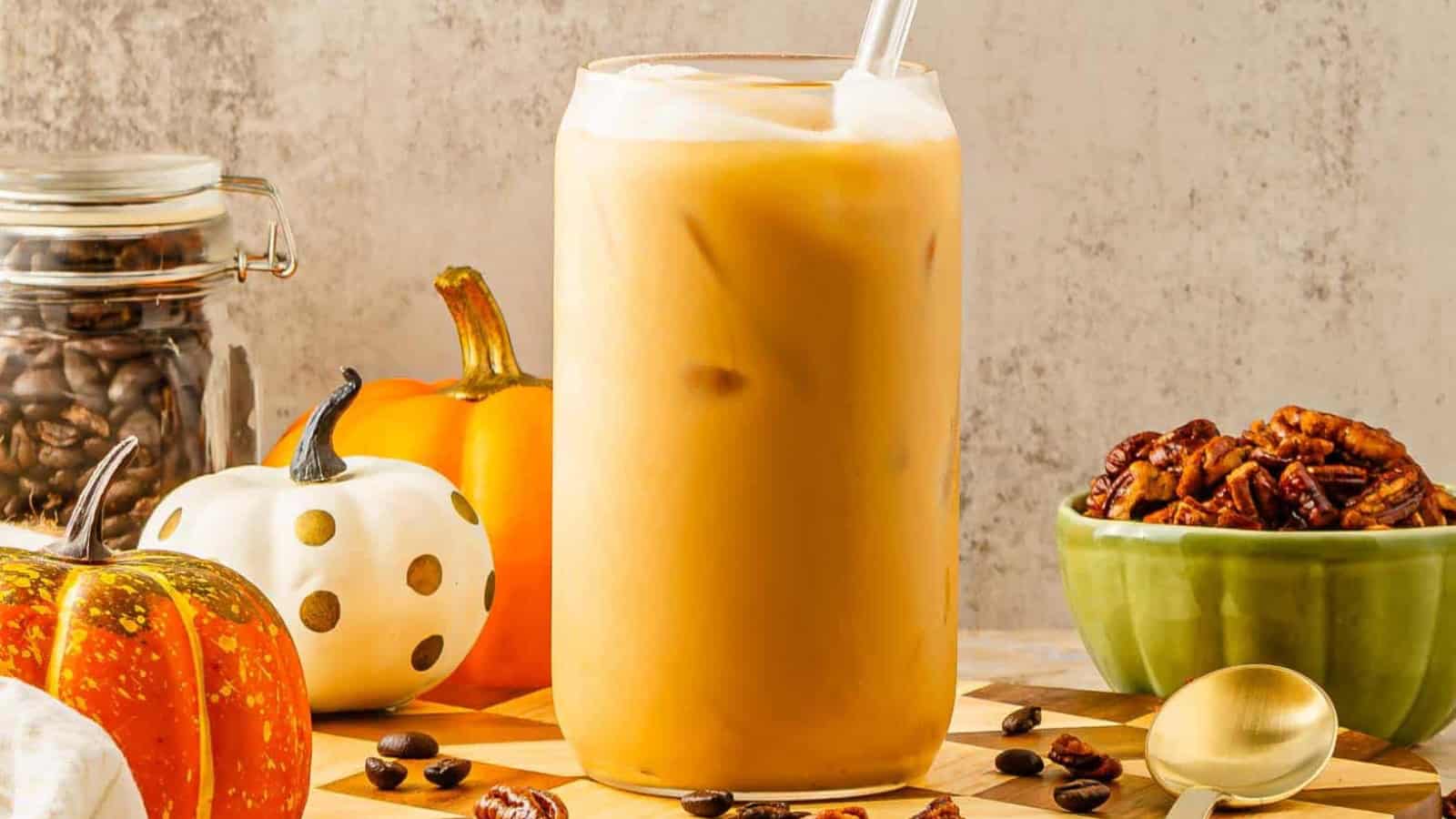 Iced coffee in a glass with pumpkins, coffee beans, and a bowl of pecans on a table.