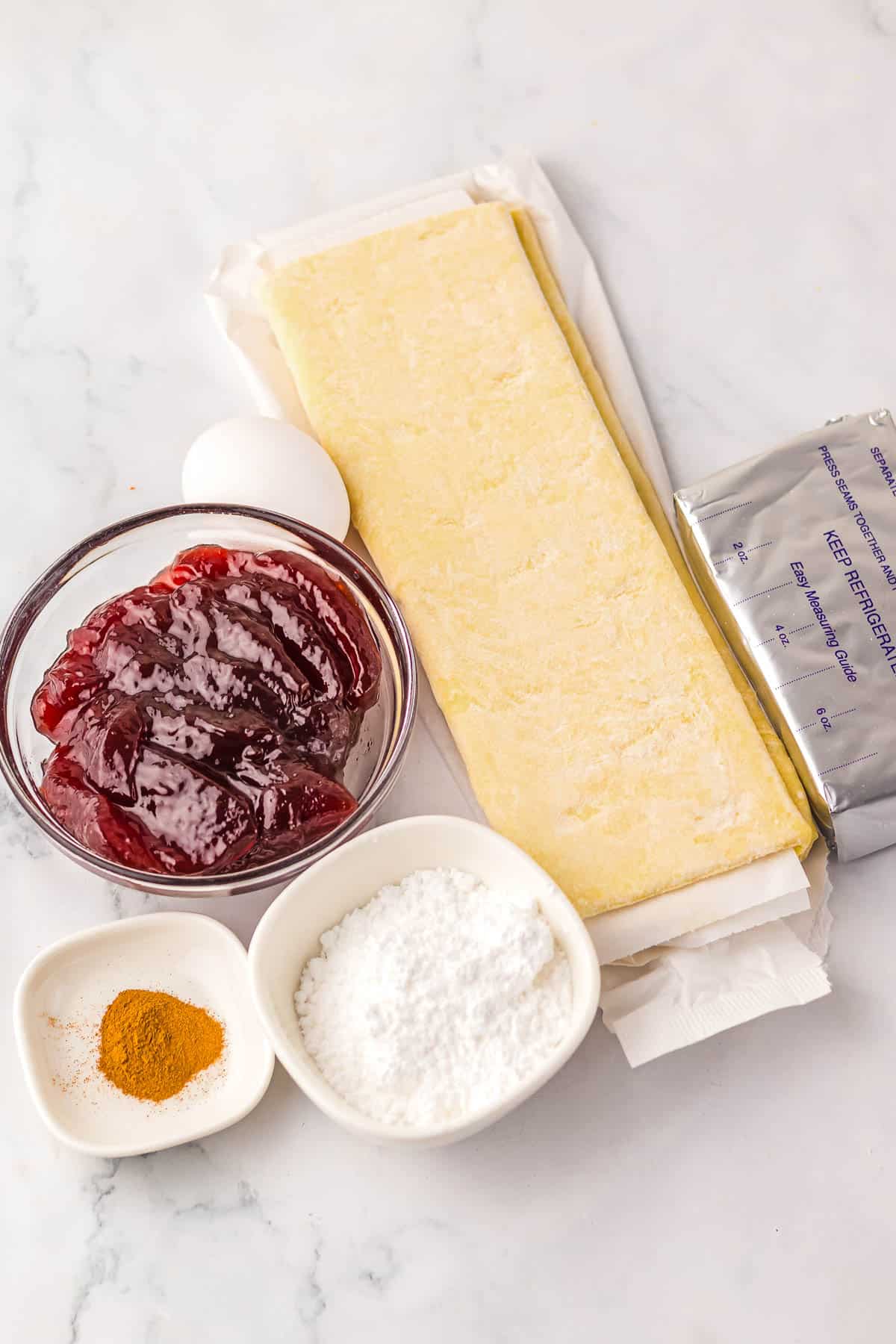 Ingredients on a counter: puff pastry, egg, cream cheese, cherry jam, powdered sugar, and spices in small bowls&mdash;everything you need to make a Starbucks Copycat Sugar Plum Cheese Danish at home.