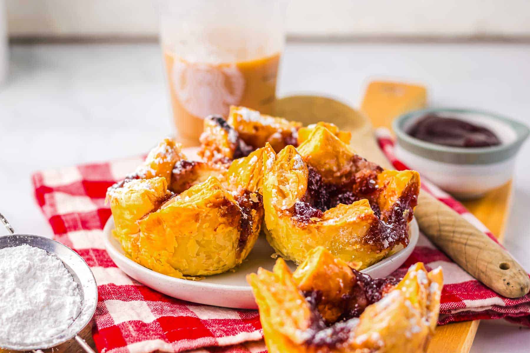 Pastries filled with jam and dusted with powdered sugar on a plate, reminiscent of a Starbucks copycat cheese danish, with iced coffee and utensils in the background.