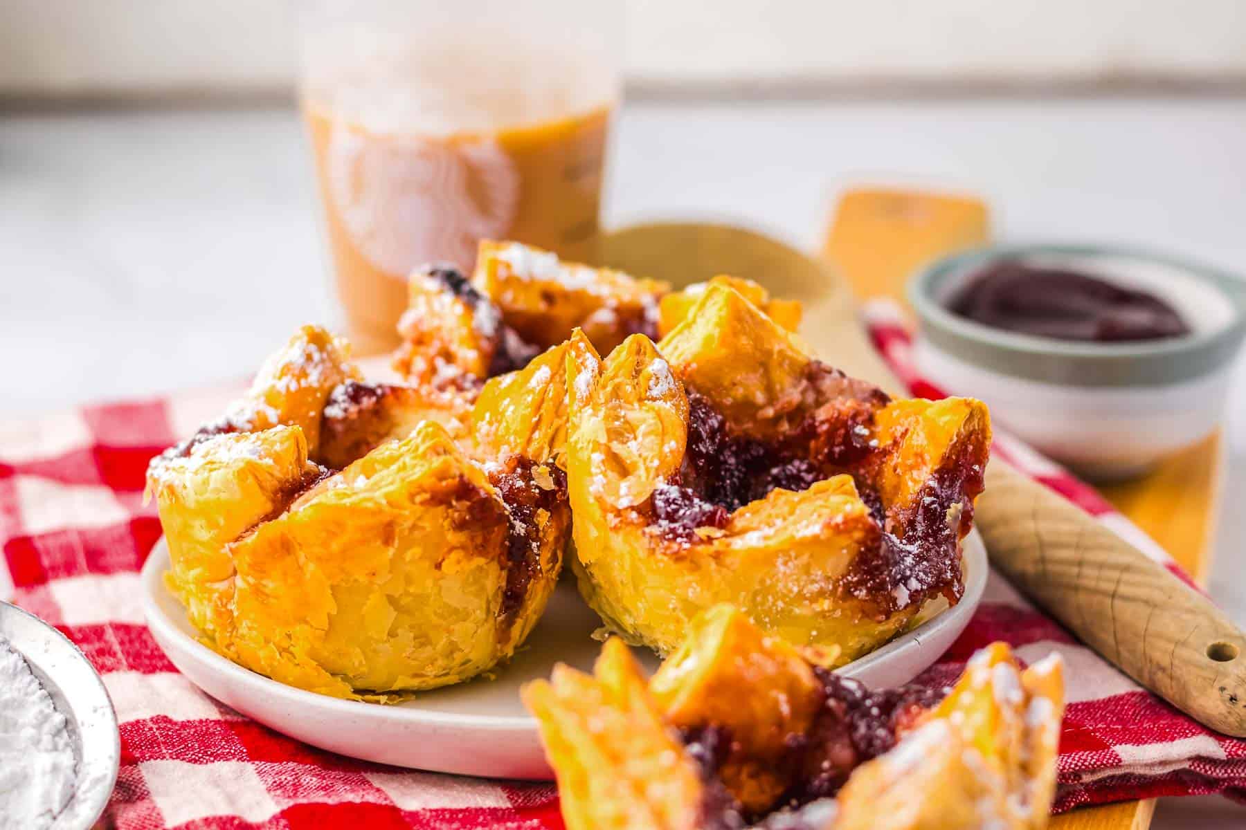 Flaky pastries with jam filling, inspired by a Starbucks copycat Sugar Plum Cheese Danish, are served on a white plate atop a red-checkered cloth, with iced coffee in the background.
