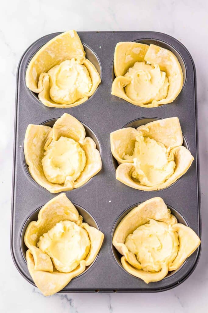A muffin tin with six unbaked pastry cups filled with cream cheese mixture, perfect for making a Starbucks copycat cheese danish, sits on a marble surface.