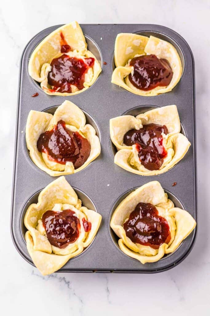 A muffin tin with six unbaked Sugar Plum Cheese Danish pastry cups filled with red jam and cream cheese on a marble surface.