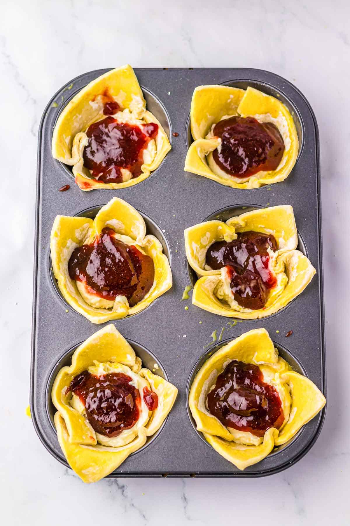 A muffin tin with six pastry cups filled with cheese and topped with red jam, these sugar plum cheese danish treats are a delicious Starbucks copycat, ready to bake.