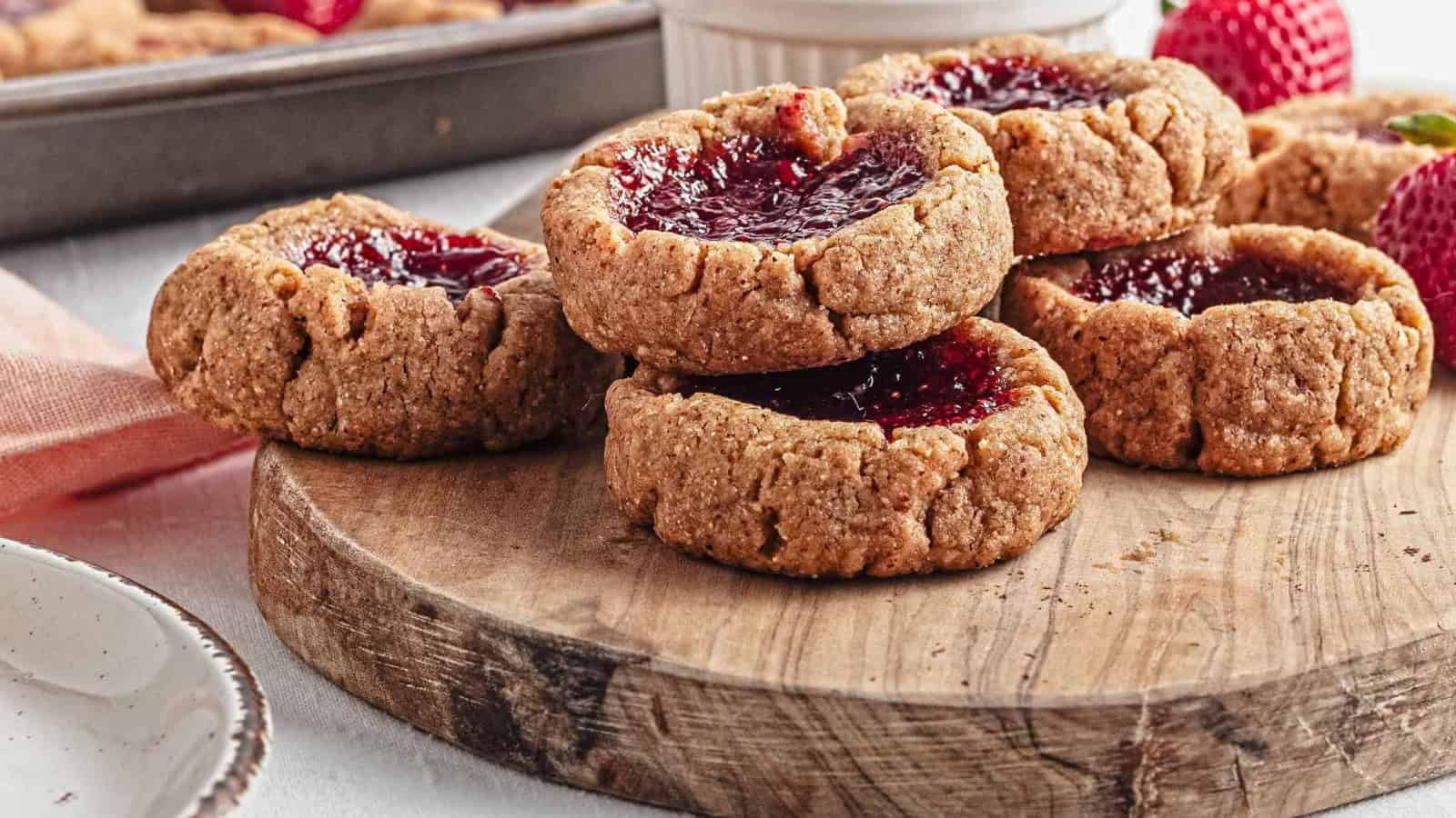 Thumbprint cookies with strawberry jam on a baking tray, surrounded by fresh strawberries.