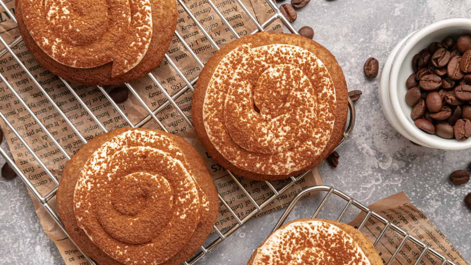 A frosted cookie dusted with cocoa powder, surrounded by coffee beans and baked cookies on a cooling rack.