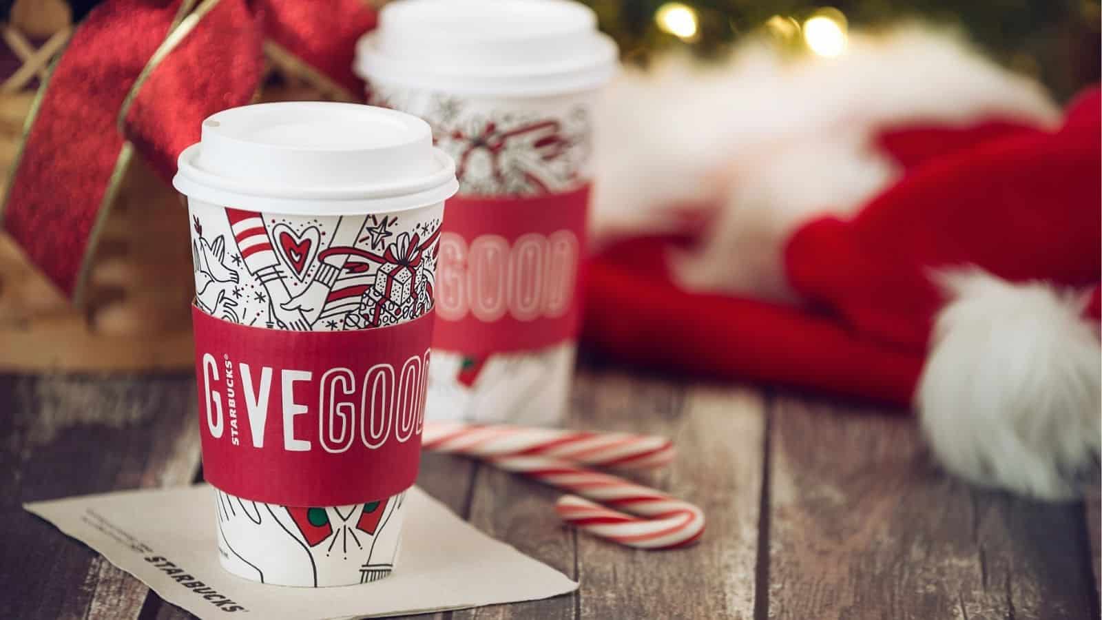Two festive Starbucks cups&mdash;perfect for hot and iced Starbucks Holiday Drinks&mdash;with "Give Good" sleeves, candy canes, and holiday decorations on a wooden table.