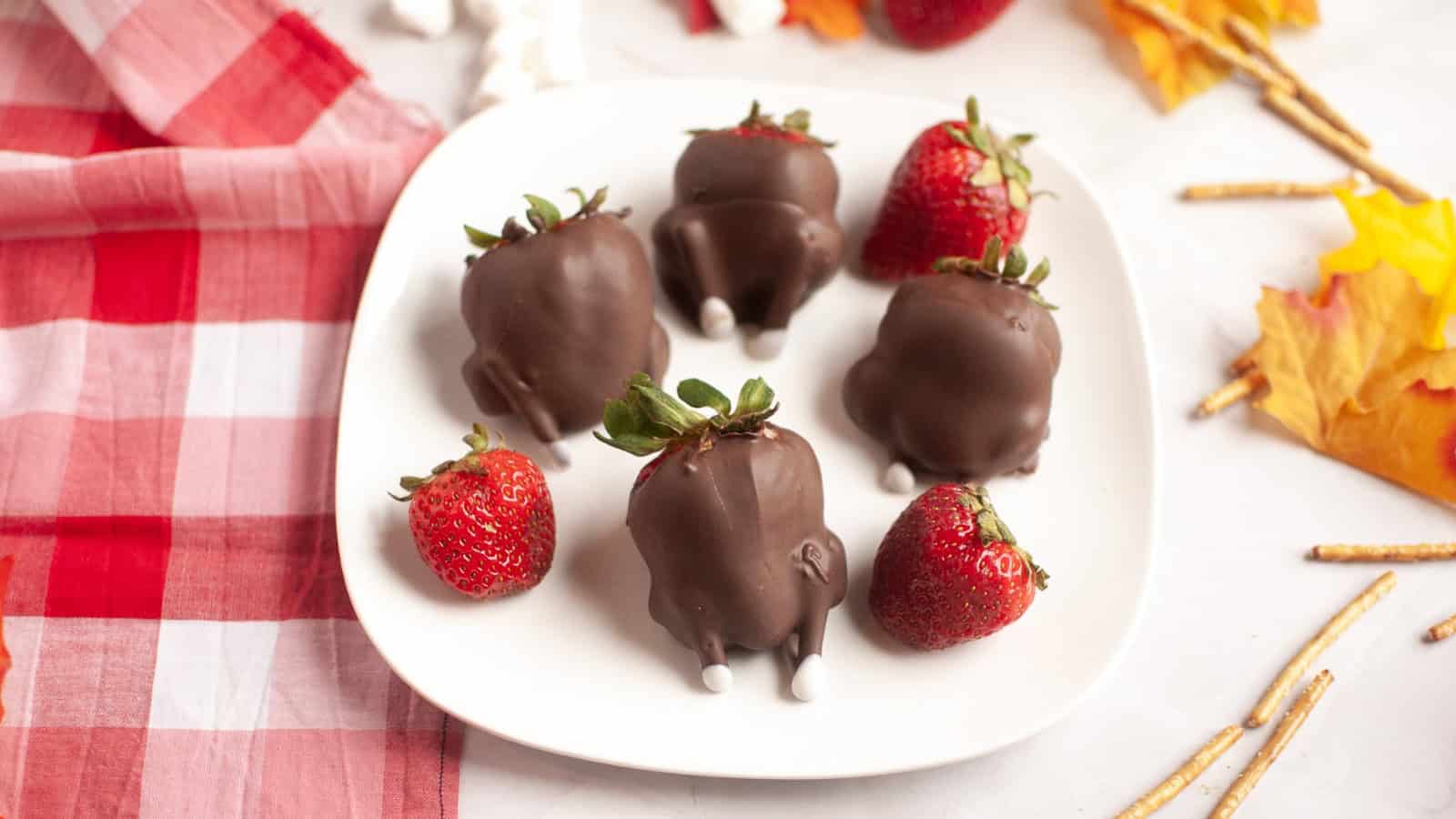 Strawberries dipped in chocolate to look like turkeys on a white plate, with fall decor and a red napkin.