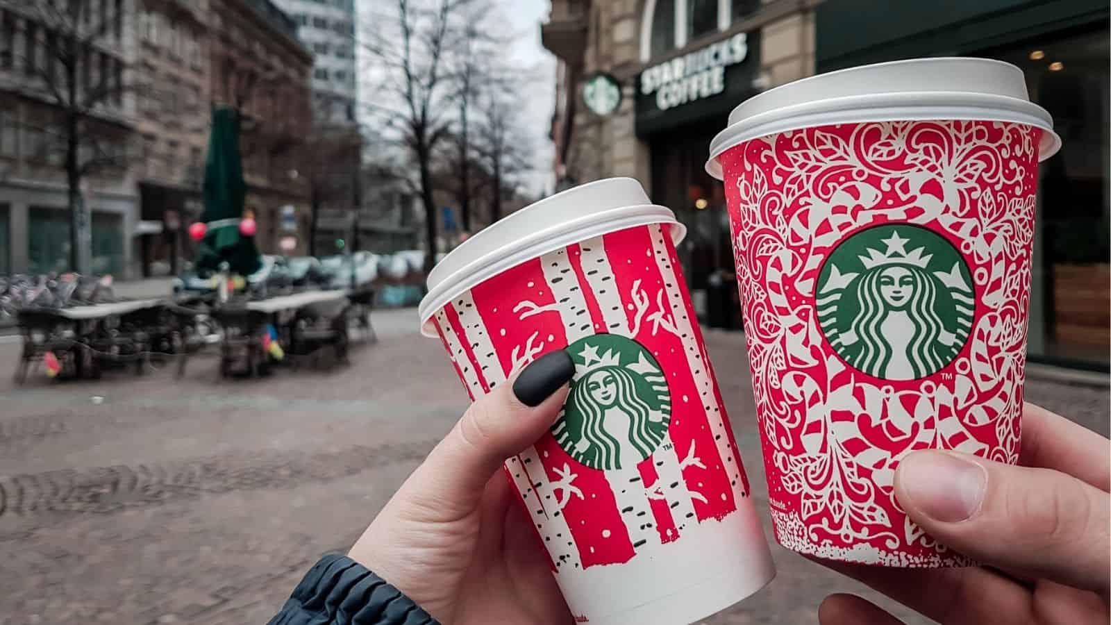 Two hands holding festive Starbucks holiday drinks outside a Starbucks store on a city street.
