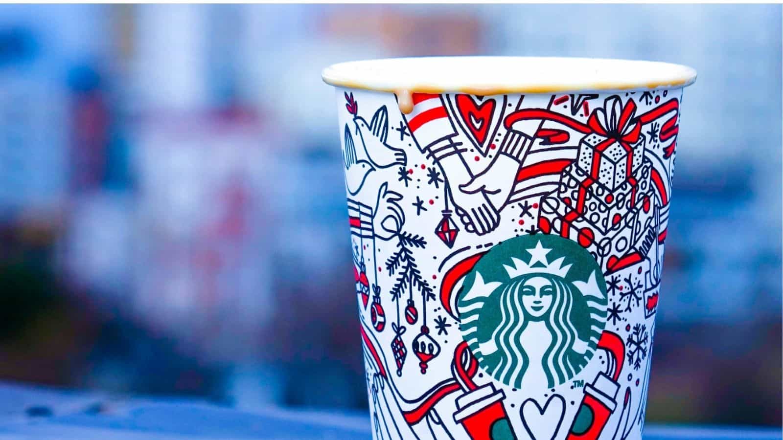 A colorful holiday-themed Starbucks cup with festive designs and the Starbucks logo, perfect for enjoying hot and iced Starbucks holiday drinks, against a blurred background.