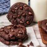 Chocolate chip cookies inspired by Crumbl copycat cookies, topped with extra chocolate chips, sit next to glasses of milk on a wooden table.