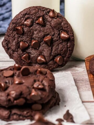 Chocolate chip cookies inspired by Crumbl copycat cookies, topped with extra chocolate chips, sit next to glasses of milk on a wooden table.