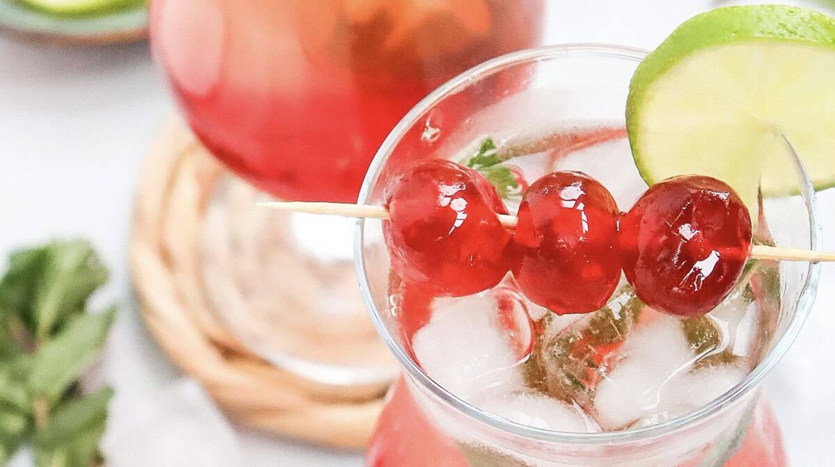 A refreshing Shirley Temple mocktail served in a glass of iced red drink, garnished with three cherries on a skewer and a lime slice. Perfect for any occasion, this recipe delights with its vibrant color and classic charm.