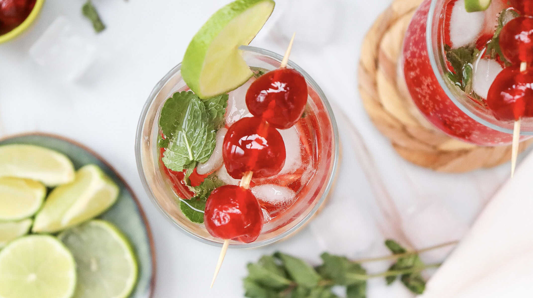 Glass of iced red Shirley Temple Mocktail with cherries, lime wedge, and mint, viewed from above. Sliced limes nearby.