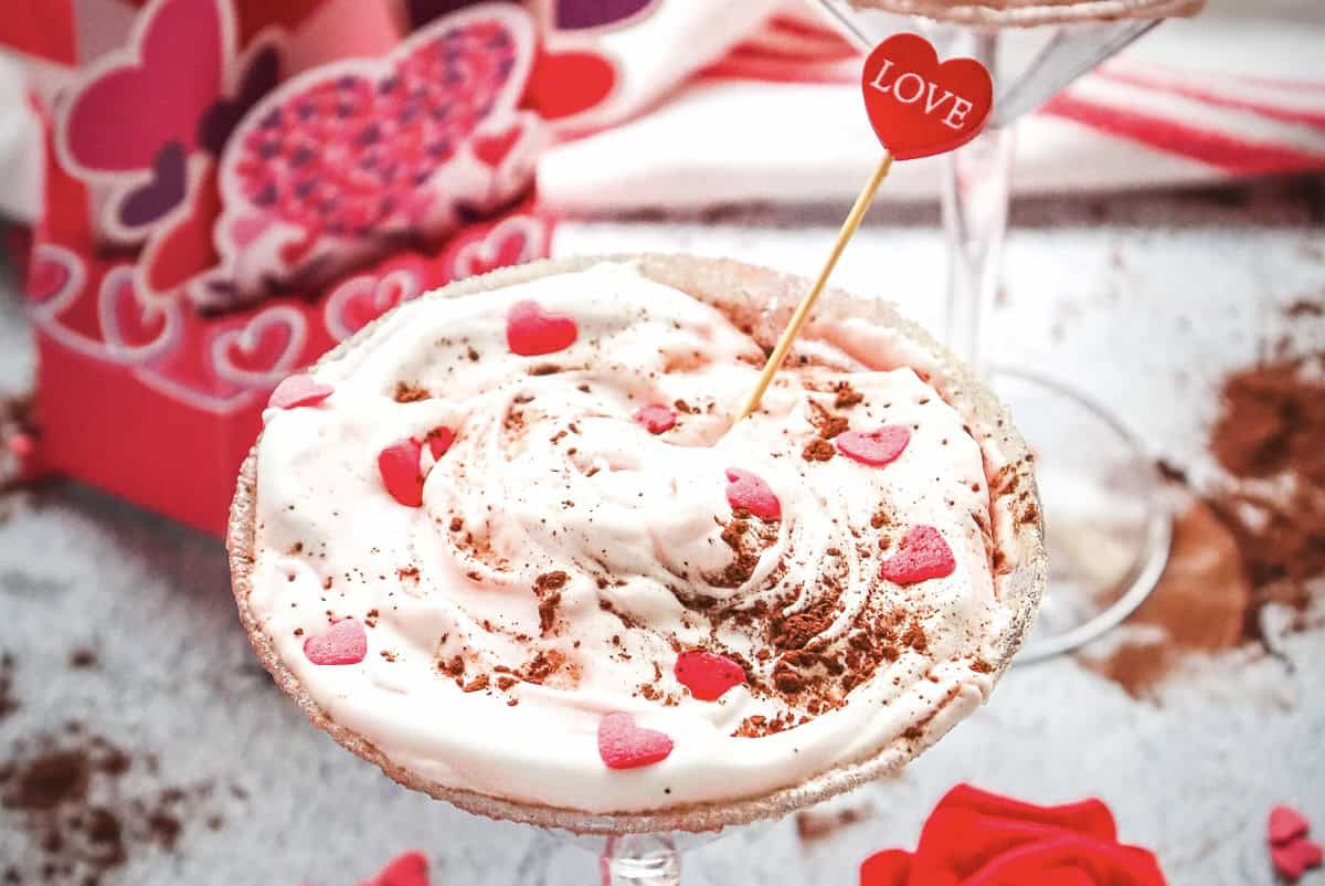 A creamy Red Velvet Chocolate Martini mocktail dessert in a glass, topped with heart sprinkles and a "LOVE" pick, set against a romantic background.