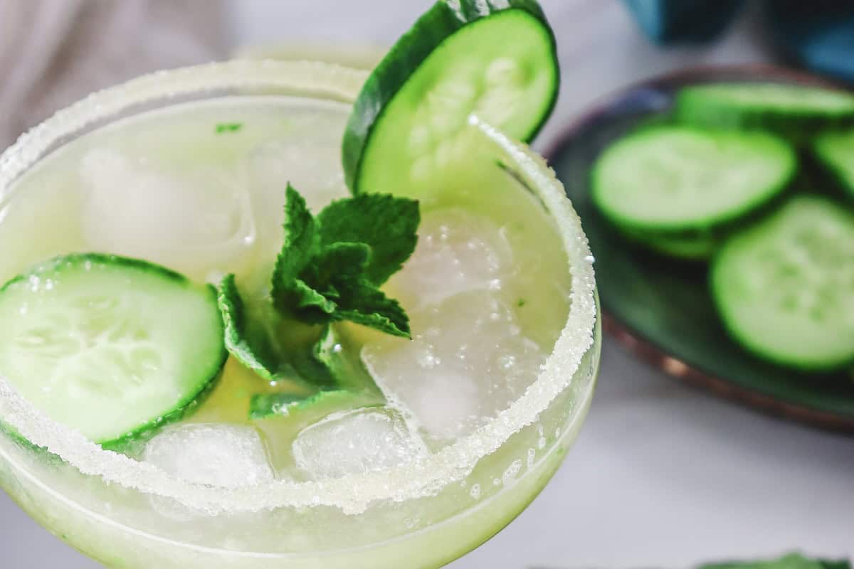 A refreshing Margarita Mocktail with ice, cucumber slices, mint, and a sugared rim, featuring extra cucumber slices in the background.