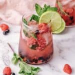A refreshing berry mojito with lime and mint, garnished with ice, raspberries, blueberries, and fresh mint on a marble surface.