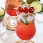 A vibrant Shirley Temple mocktail, this red iced cocktail is garnished with cherries and a lime slice, served in a curvy glass on a coaster.