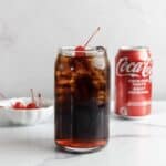 A glass of cola with ice and cherries, a bowl of cherries, and a Coca-Cola can on a white surface evoke the classic Roy Rogers&mdash;a beloved non-alcoholic cocktail inspired by the vintage drink recipe.