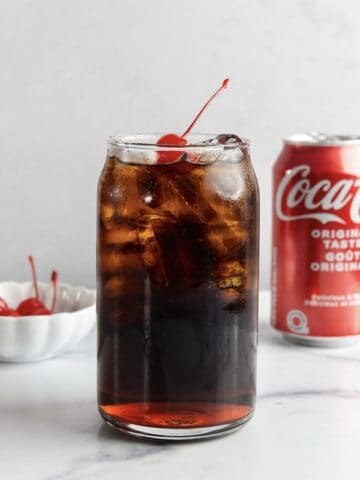 A glass of cola with ice and cherries, a bowl of cherries, and a Coca-Cola can on a white surface evoke the classic Roy Rogers—a beloved non-alcoholic cocktail inspired by the vintage drink recipe.