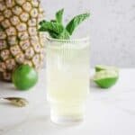 A tall glass of Tropical Electrolyte Mocktail garnished with mint, with a pineapple and lime in the background.