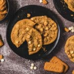 A black plate with three white chocolate chip cookies, one broken, surrounded by biscuits and white chips&mdash;these treats are perfect for any Biscoff cookies or Crumbl copycat fan.