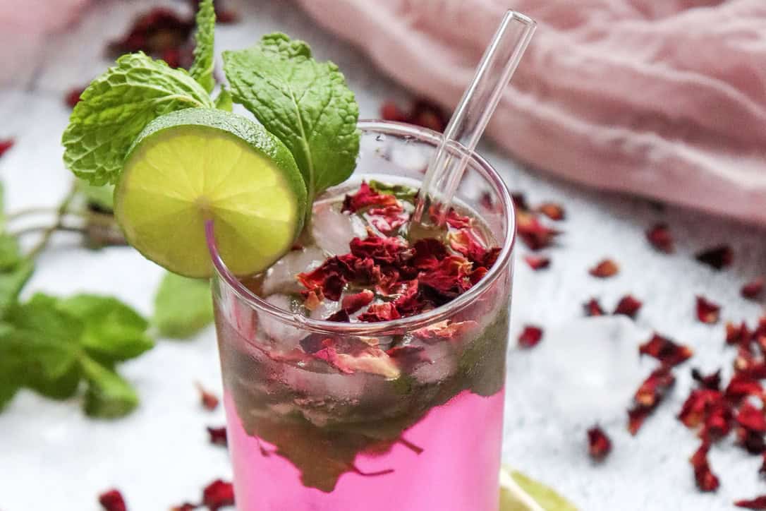 A refreshing non-alcoholic rose mojito mocktail recipe: a pink drink with ice, mint, lime slice, dried petals, and a glass straw, garnished with fresh mint.