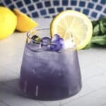 A vibrant Purple Mocktail topped with a lemon slice and purple flower, set against a backdrop of lemons and mint.