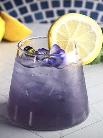 A vibrant Purple Mocktail topped with a lemon slice and purple flower, set against a backdrop of lemons and mint.