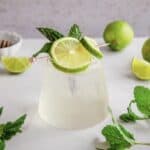 A refreshing mocktail garnished with lime slices and mint, surrounded by fresh limes and mint leaves on a white surface&mdash;perfect for trying a new recipe.