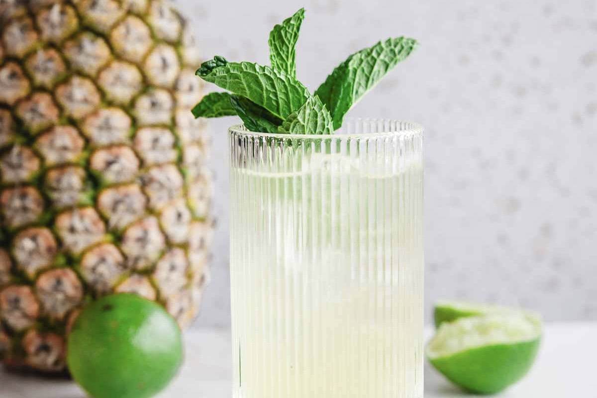 A glass of iced lime drink with mint leaves, next to a pineapple and halved lime&mdash;perfect as a refreshing Tropical Electrolyte Mocktail on a sunny day.