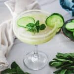 A cocktail glass with a refreshing Cucumber Margarita Mocktail, ice, mint sprig, and cucumber slices, on a light surface.