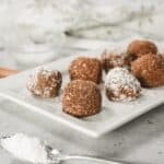 Cream Cheese Truffles dusted with powdered sugar rest on a white plate, with a bowl of powdered sugar in the background.