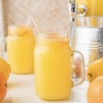 Glass jars filled with homemade orangeade and a glass straw, surrounded by fresh oranges and lemons.