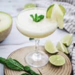 A frosty Melon Margarita cocktail garnished with mint, lime slice, and melon, served on a wooden board with extra lime and mint.