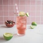 A pink pomegranate mocktail with ice, lime slices, and a striped straw, served on a marble surface alongside fresh lime and pomegranate&mdash;perfect for an electrolyte boost.