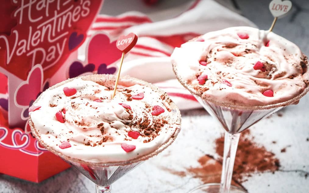 Two pink creamy desserts in martini glasses, inspired by a Red Velvet mocktail recipe, topped with heart sprinkles and "love" picks, surrounded by Valentine&rsquo;s decor.