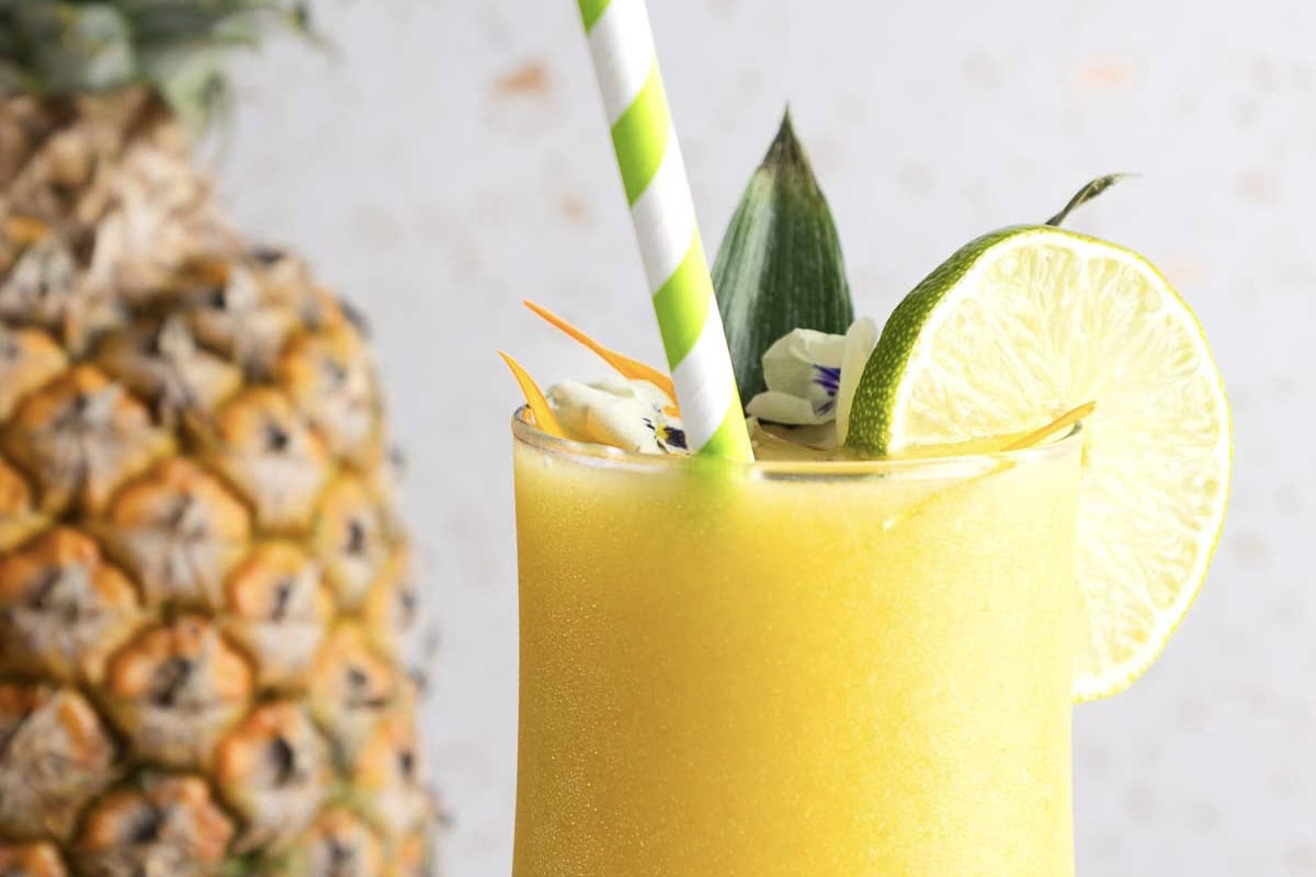 Yellow smoothie in a glass with a green striped straw, lime slice, and pineapple&mdash;this vibrant Mango Colada mocktail recipe shines with tropical flavors, complete with a whole pineapple in the background.