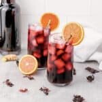 Two glasses of iced hibiscus tea, inspired by a classic Agua de Jamaica recipe, with orange slices, surrounded by hibiscus petals and a bottle in the background.
