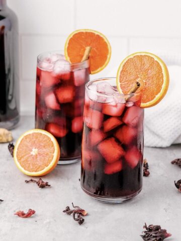Two glasses of iced hibiscus tea, inspired by a classic Agua de Jamaica recipe, with orange slices, surrounded by hibiscus petals and a bottle in the background.