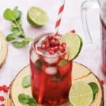 A glass of Virgin Pomegranate iced drink with pomegranate seeds, lime slice, mint leaves, and a red-striped straw.