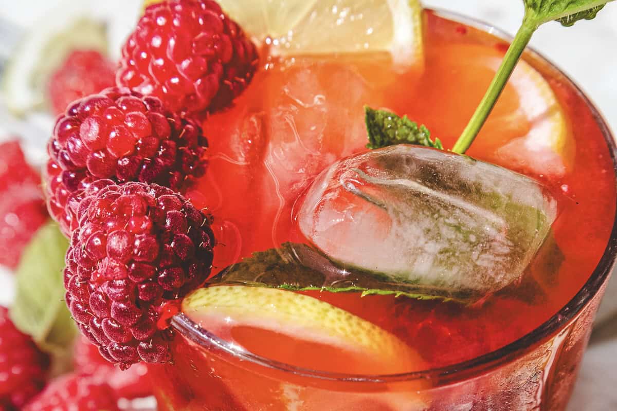 A close-up of a refreshing raspberry mojito mocktail recipe with raspberries, lemon slices, mint, and ice cubes.