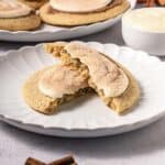 A frosted cinnamon swirl cookie is broken in half on a white plate, with more delicious Crumbl copycat cookies in the background.