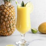 Tropical yellow Mango Colada mocktail in a glass with lime slice, straws, and pineapple garnish; pineapple and mango nearby.