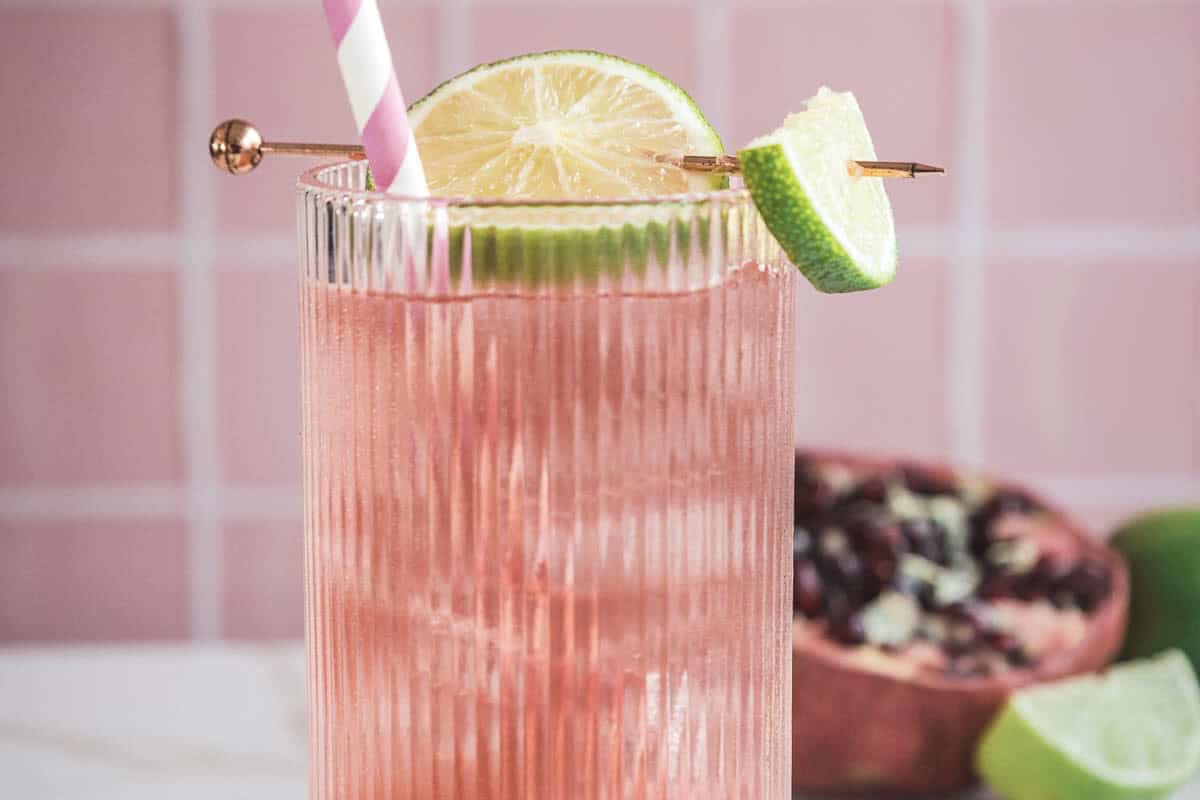 A refreshing pink mocktail recipe with ice, lime slices, and a striped straw in a ribbed glass, featuring pomegranate in the background for a boost of natural electrolytes.