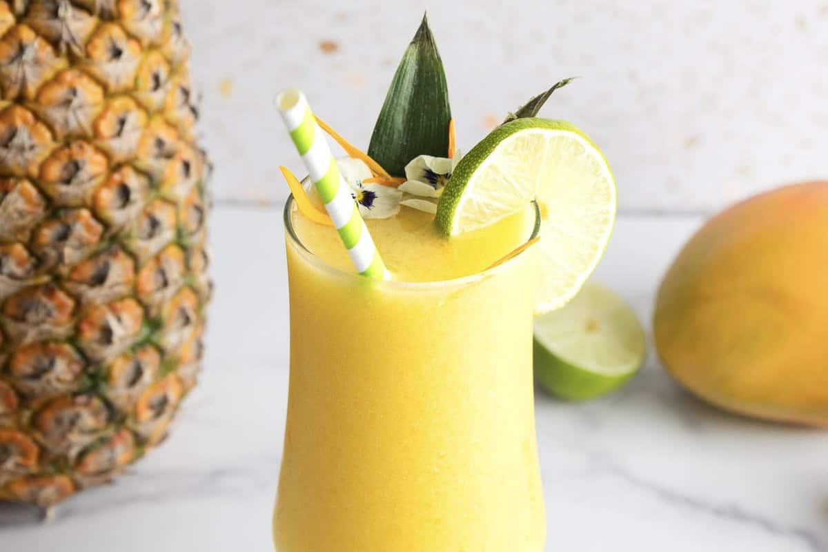 A tropical Mango Mocktail garnished with a lime slice, pineapple leaf, and striped straws, with fresh fruits in the background&mdash;perfect for anyone seeking a refreshing mocktail recipe.