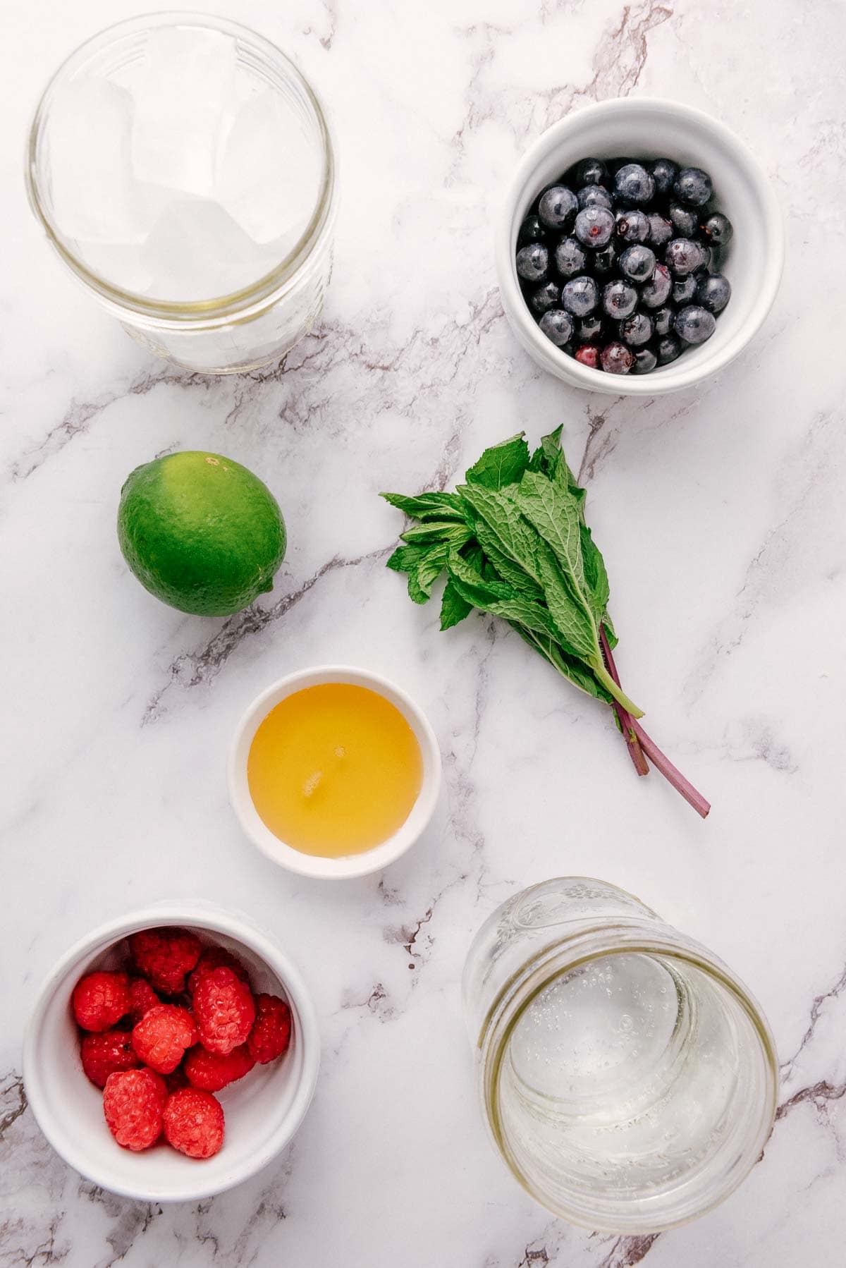 A flat lay of blueberries, raspberries, lime, mint, honey, ice cubes, and a jar of water on a marble surface&mdash;perfect ingredients for a refreshing Blueberry Mint Mocktail.