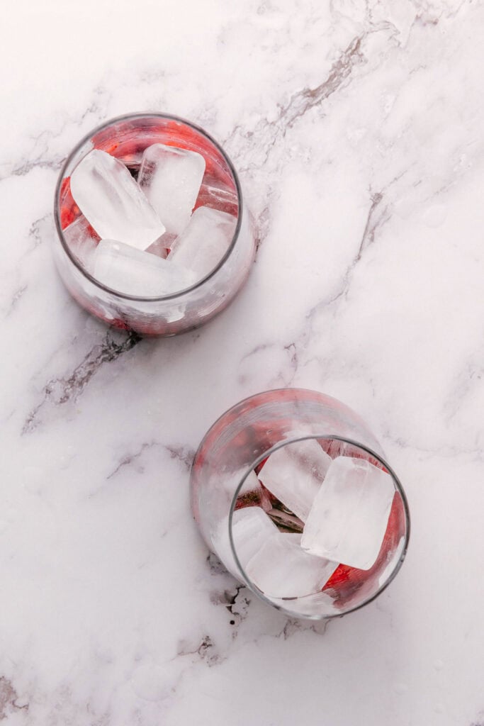 Two glasses with ice cubes and a refreshing raspberry mint mocktail, placed on a white marble surface.