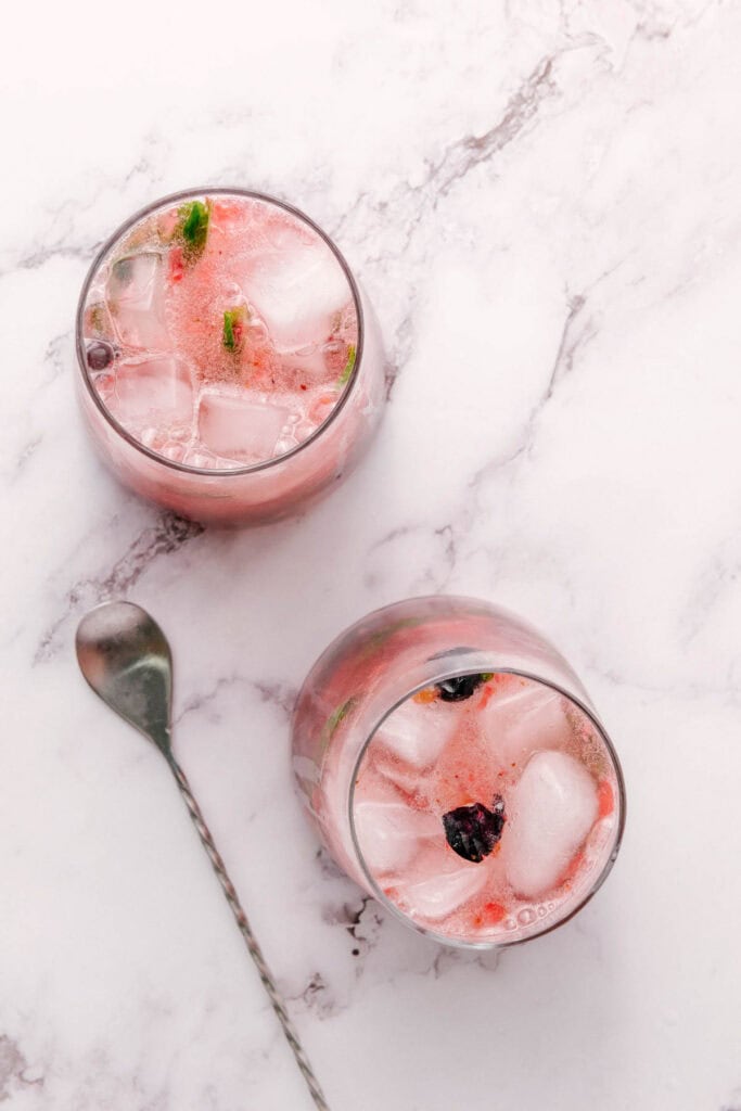 Two glasses of mint mocktail with pink iced drinks, garnished with raspberry, blueberry, and fresh mint on a marble surface, next to a metal stirring spoon.